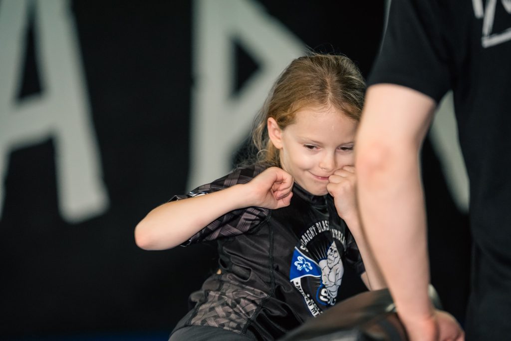Smiling girl in an SBG rash guard practising strikes on a coach's pad