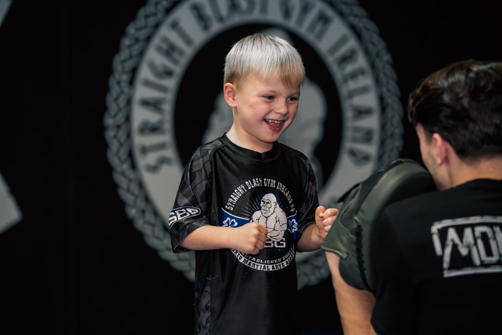 Smiling boy in an SBG rash guard hitting pads with a coach